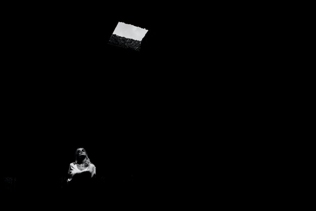 Black and white photo of a woman in a black room looking up at a single window of light.