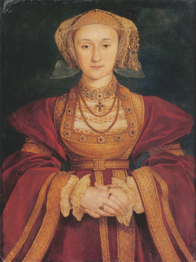 A painting of a woman in a gold and red velvet gown with ornate embroidery, an elaborate gold cap and veil, and lots of jewelry. She is peaceful, with pale skin, dark eyes, no hair visible under her cap, and her hands folded demurely in her lap. Portrait by Hans Holbein the Younger, 1539.
