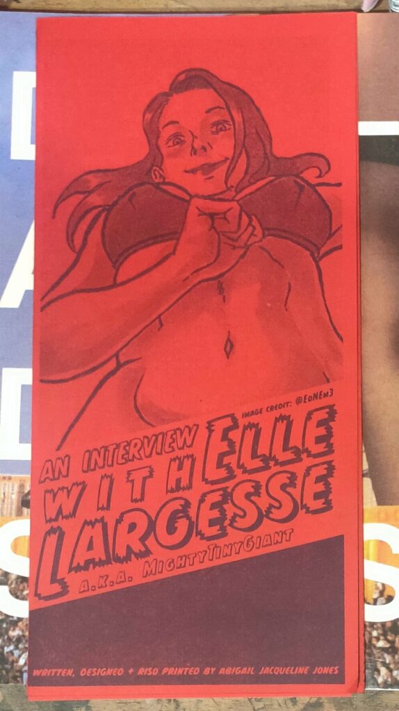 A red zine printed with a low-angle illustration of Elle Largesse in a bra and panties, drawn by @EdNEm. The title in block letters reads An Interview With Elle Largesse AKA MightyTinyGiant. Written, Designed, and Riso-Printed by Abigail Jaqueline Jones.