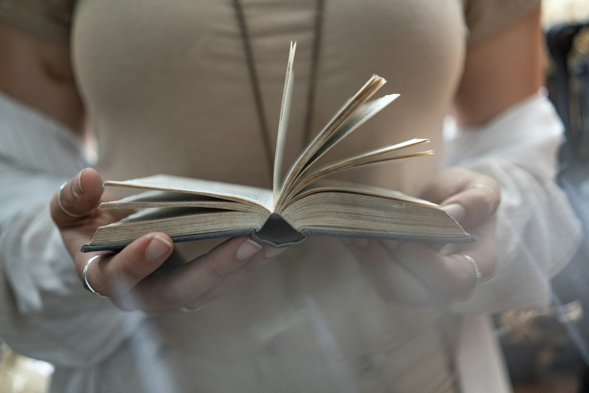 A low-angle photo of a woman holding a book and allowing the pages to fall open like a fan. She's wearing silver rings, a long pendant, a tan tank top, and there's some kind of white shirt or sweater sliding down her arms to bare a tattoo that's too blurry to make out. Image credit to Jessica Da Rosa on Unsplash.